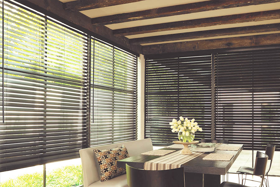You don't have to worry about broken blinds on sunroom windows when you install high-quality wood blinds! Dark wood window blinds on windows in a sunroom with a dining table
